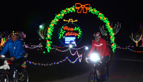 Two cyclists ride through a festive, illuminated park entrance with a "Happy Holidays" sign and colorful lights.