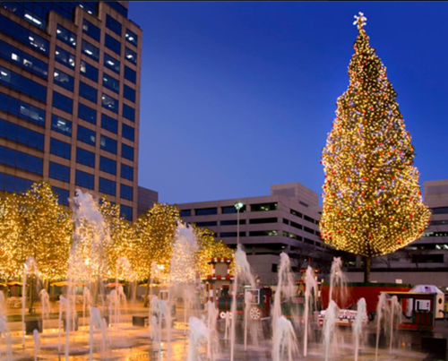A brightly lit Christmas tree stands in a plaza, surrounded by illuminated trees and a fountain, against a twilight sky.