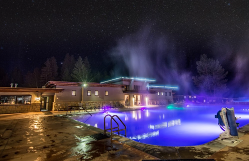 A serene outdoor pool at night, illuminated with colorful lights under a starry sky, surrounded by trees.