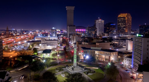 A nighttime cityscape featuring illuminated buildings and a monument in the foreground, with traffic lights in the background.