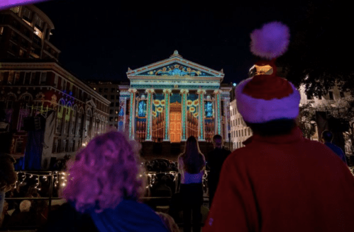People watch a colorful light projection on a historic building during a nighttime event.