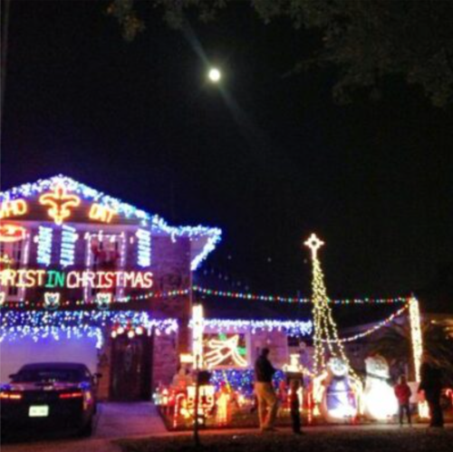 A house decorated with colorful Christmas lights, a large snowman, and a tree under a bright moon at night.