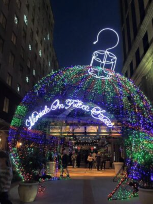 A festive archway shaped like a Christmas ornament, adorned with colorful lights, welcoming visitors to a holiday event.