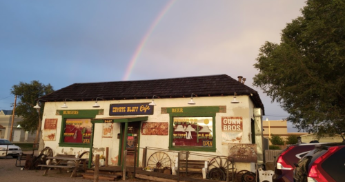 A rustic cafe with a rainbow overhead, featuring signs for burgers and beer, surrounded by vintage decor.