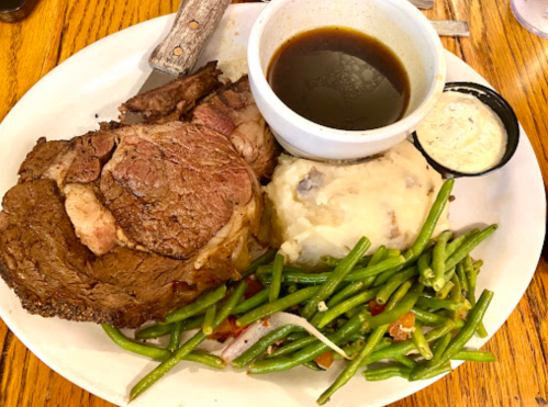 A plate featuring a large slice of prime rib, mashed potatoes, green beans, and a bowl of gravy.