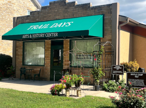 A building with a green awning labeled "Trail Days Arts & History Center," surrounded by flowers and signage.