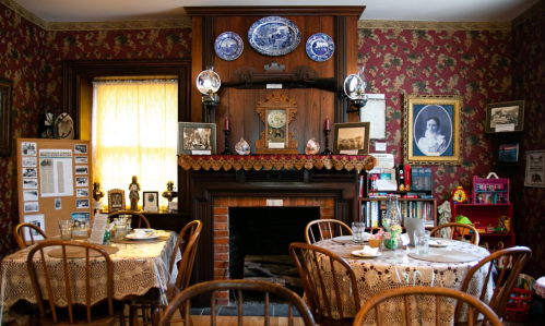 Cozy dining room with wooden tables, floral wallpaper, a fireplace, and vintage decor, creating a warm, inviting atmosphere.