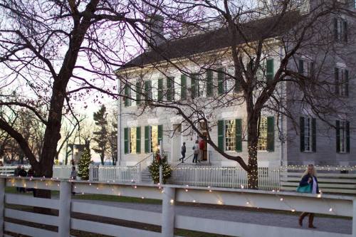 A historic building with green shutters, surrounded by trees and festive lights, as people walk nearby at dusk.