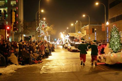 Two elves walk down a festive street lined with holiday lights and a crowd watching a nighttime parade.