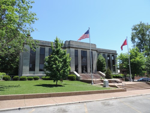 A modern courthouse building with large windows, surrounded by trees and flags, on a sunny day.
