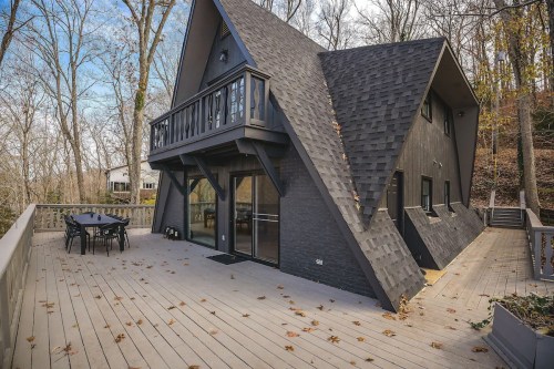 A modern A-frame house with a dark exterior, surrounded by trees and featuring a spacious wooden deck.