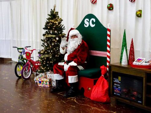 Santa Claus waves from a festive chair beside a decorated Christmas tree and gifts, including a bicycle and stockings.