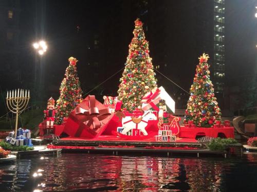 Colorful Christmas trees and decorations by a pond, featuring nutcrackers, gifts, and a menorah in a festive display.