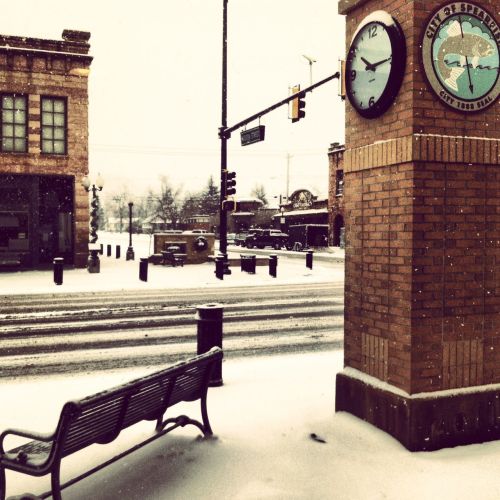 A snowy street scene featuring a clock tower, a bench, and a traffic light in a quaint town.