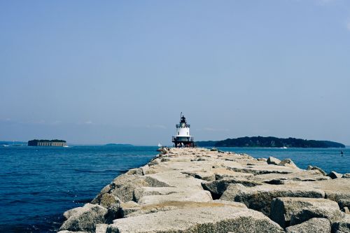 A lighthouse stands at the end of a rocky jetty, with calm blue waters and distant land visible in the background.