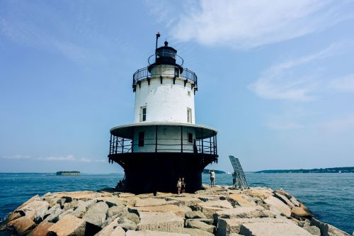 A white lighthouse stands on a rocky pier, surrounded by water under a clear blue sky.