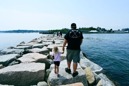 A person and a child walk hand-in-hand along a rocky jetty by the water on a sunny day.