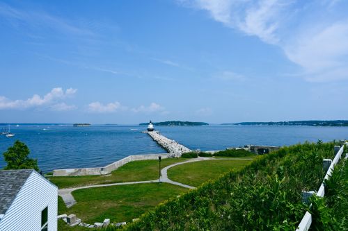 A scenic view of a lighthouse on a pier, surrounded by calm waters and a clear blue sky. Green grass in the foreground.