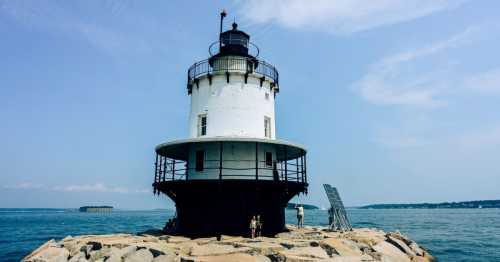 A white lighthouse stands on a rocky jetty, surrounded by calm water under a clear blue sky.