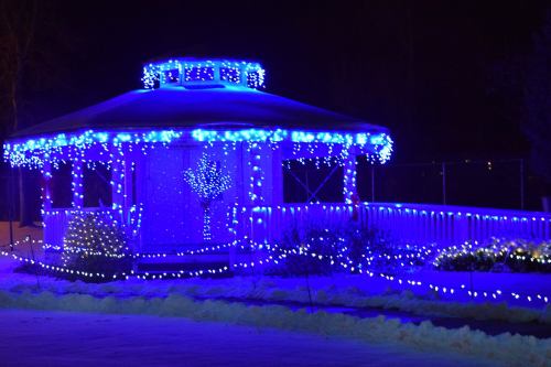 A gazebo adorned with blue and white lights, surrounded by snow, creating a festive nighttime scene.