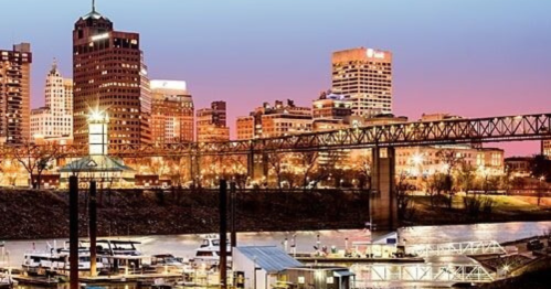 City skyline at dusk with illuminated buildings and a bridge over a river, featuring boats in the foreground.