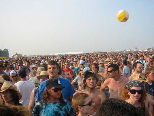 A large crowd of people at an outdoor festival, with some wearing hats and a beach ball flying through the air.