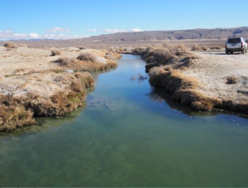 A narrow, winding stream flows through a dry landscape with sparse vegetation and a distant vehicle parked nearby.