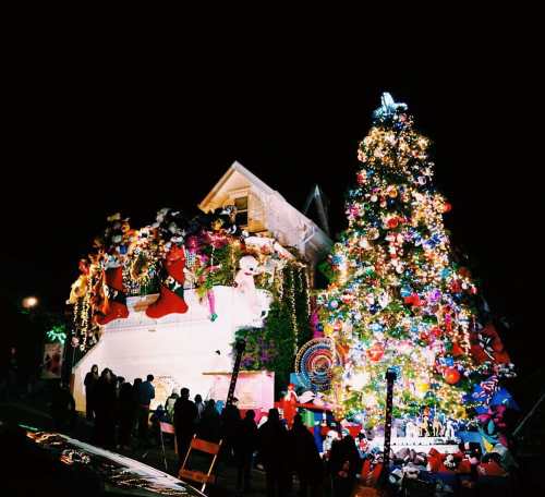 A brightly lit house decorated for Christmas, featuring a large tree and festive ornaments, with people admiring the display.