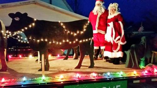 Santa and Mrs. Claus stand beside a decorated cow on a festive float, illuminated by colorful lights at night.