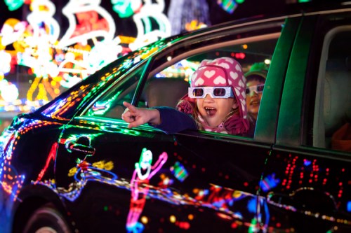 A child in heart-patterned glasses excitedly points out colorful holiday lights from a car window.