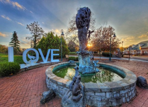 A park scene featuring a fountain with a statue, surrounded by greenery and large letters spelling "LOVE" at sunset.
