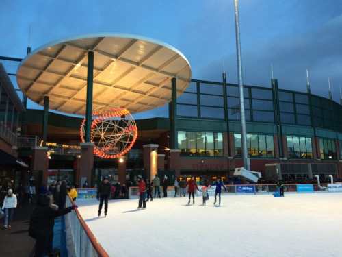 Outdoor ice skating rink with people skating, surrounded by a modern building and a large circular structure overhead.