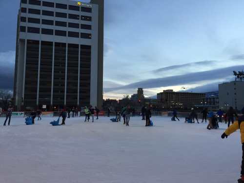 People ice skating on a rink with a city skyline in the background during twilight.