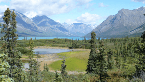 A scenic view of mountains and a lake surrounded by lush greenery and trees under a partly cloudy sky.