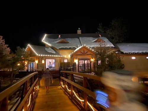 A cozy, illuminated lodge at night, with a wooden bridge leading to its entrance and snow on the roof.