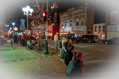A nighttime street scene with people sitting along the sidewalk, festive lights, and vintage buildings in the background.