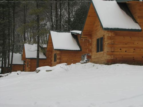 A row of wooden cabins covered in snow, surrounded by trees in a winter landscape.