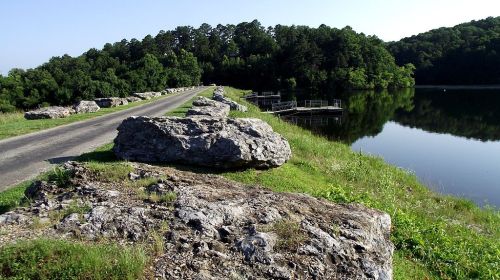 A serene lakeside view with a rocky path, lush greenery, and calm water reflecting the trees.