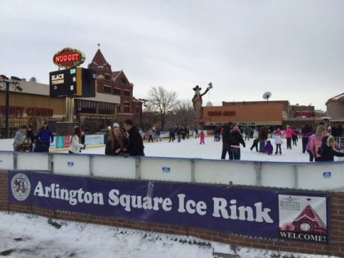 People ice skating at Arlington Square Ice Rink, with a welcoming sign and a nearby casino in the background.