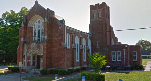 A brick church building with a tall tower, surrounded by trees and a grassy area, featuring a clear blue sky.