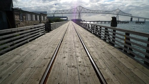 A wooden pier extends over water, leading to a bridge in the distance under a cloudy sky.