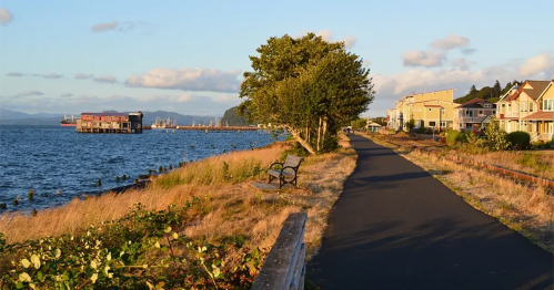 A scenic waterfront path lined with trees, benches, and houses, with a view of the water and distant mountains.