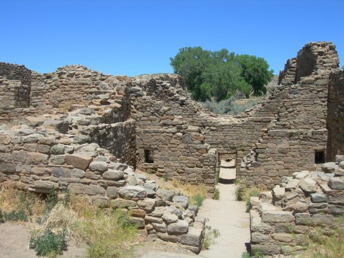 Ancient stone ruins with a pathway, surrounded by greenery and clear blue sky.