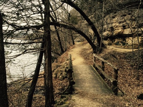 A wooden bridge crosses a path by a river, surrounded by trees and rocky cliffs in a serene, natural setting.