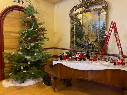 A decorated Christmas tree beside a grand piano, with festive ornaments, a mirror, and holiday figurines on display.