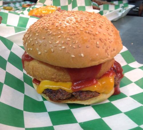 A sesame seed bun burger with cheese, ketchup, and a crispy onion ring on a green and white checkered paper.