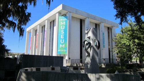 A modern building with banners reading "Library, Museum, Archives" surrounded by trees and a sculpture in front.