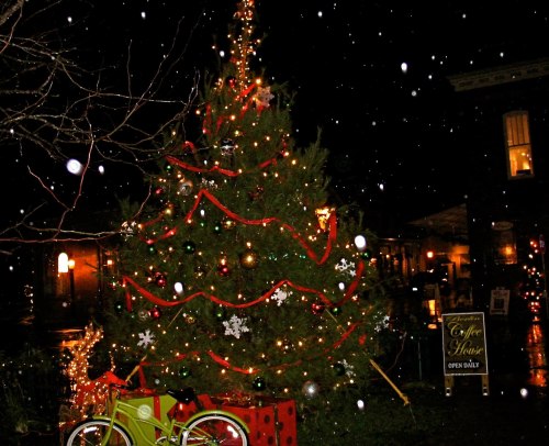 A decorated Christmas tree with lights and ornaments, surrounded by snowflakes at night. A coffee shop sign is nearby.
