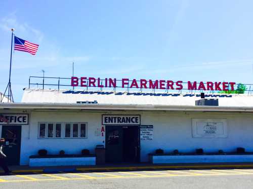 Sign for Berlin Farmers Market with an American flag, featuring an office and entrance on a sunny day.