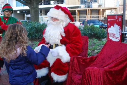 A child with curly hair interacts with Santa Claus, who is seated in a festive setting with red chairs.